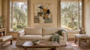 Salon lumineux d’une maison d’artistes en Provence, avec murs à la chaux beige, canapé en lin naturel, tableau abstrait contemporain, mobilier en bois patiné et vue sur des oliviers.